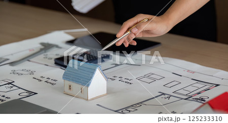 Model Building and Project Planning. A hand points to a miniature house on a blueprint, illustrating home construction planning. Model Building and Project Planning. A hand points to a miniature house on a blueprint, illustrating home construction planning. 125233310