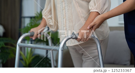 Supportive Care and Mobility Guidance. A caregiver holds the hand of an elderly patient while assisting her with a walker, enhancing safety and trust. 125233367