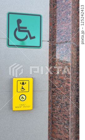 Accessibility assistance button with wheelchair symbol, braille inscription, and bell icon mounted on wall near large green sign indicating accessible entrance, promoting inclusive infrastructure Accessibility assistance button with wheelchair symbol, braille inscription, and bell icon mounted on wall near large green sign indicating accessible entrance, promoting inclusive infrastructure 125242413