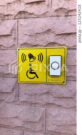 Vertical photo of bright yellow accessibility call button with tactile Braille and wheelchair icon mounted on textured pink wall, emphasizing inclusive access and support for disabled persons 125242418