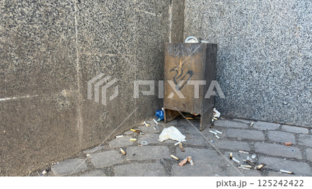 Symbol of urban fatigue and neglect overflowing smoking bin surrounded by cigarette butts on cracked pavement, visual metaphor for decay, habits, and slow erosion of public space 125242422