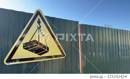 Warning sign on green fence at construction site shows crane lifting heavy load, indicating danger of falling objects under clear blue sky, symbol of safety regulation and caution 125242424