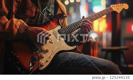 Musician playing electric guitar with ambient stage lighting. Close-up shot of hands on Stratocaster guitar strings with warm atmospheric concert lighting in background 125244110