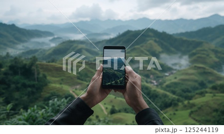 Hands holding smartphone capturing misty mountain landscape, framing green valleys and peaks through device screen. Modern travel photography concept with atmospheric scenery 125244119