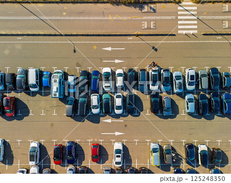 Aerial view of a large, organized parking lot filled with colorful cars. Bright sunlight casts long shadows, highlighting the neat rows and road markings surrounding the area. Aerial view of a large, organized parking lot filled with colorful cars. Bright sunlight casts long shadows, highlighting the neat rows and road markings surrounding the area. 125248350