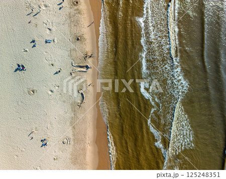 Aerial view of a beach where gentle waves meet the sandy shore. People are scattered across the sand, some building sandcastles, others relaxing and enjoying the sunny day. 125248351
