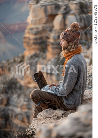 Man during hike sitting on a rock with a laptop on his lap. Remote work. Sitting on a rock, the man proves that the great outdoors can serve as a perfect setting for remote work. 125248978