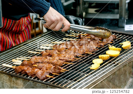 Chef grilling meat skewers and potatoes on barbecue. Hand holds tongs preparing traditional kebabs on street food festival. Outdoor cooking process. Chef grilling meat skewers and potatoes on barbecue. Hand holds tongs preparing traditional kebabs on street food festival. Outdoor cooking process. 125254782