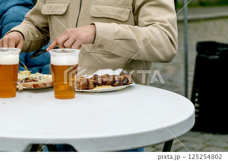 Person enjoying grilled meat skewers and draft beer at outdoor table. Casual dining scene with person in beige jacket eating street food. 125254802