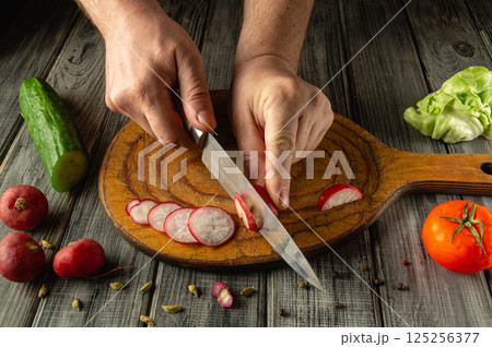 Hands expertly slice radishes on a wooden board surrounded by fresh vegetables like cucumber, tomato, and lettuce in a cozy kitchen ambiance 125256377