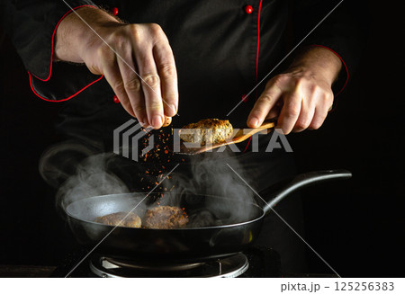 A chef skillfully adds spices to sizzling meat patties in a frying pan, producing flavorful aromas in a kitchen filled with steam during a nighttime cooking session A chef skillfully adds spices to sizzling meat patties in a frying pan, producing flavorful aromas in a kitchen filled with steam during a nighttime cooking session 125256383