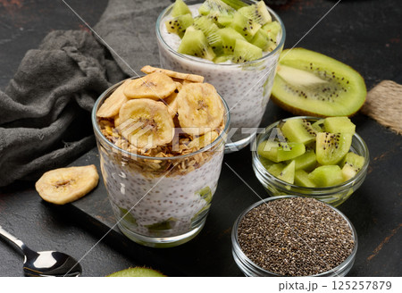 Two glasses of chia seed pudding, one topped with diced kiwi and the other with granola and banana chips, are on a dark surface with ingredients Two glasses of chia seed pudding, one topped with diced kiwi and the other with granola and banana chips, are on a dark surface with ingredients 125257879