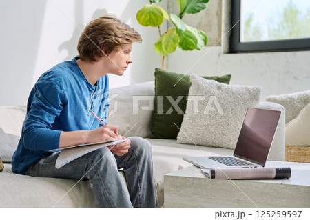 Teenager guy student studying sitting at home on sofa using laptop textbook notebook 125259597