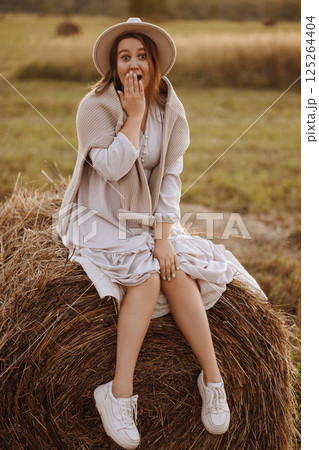 Young woman in beige dress sitting on haystack in village field Young woman in beige dress sitting on haystack in village field 125264404