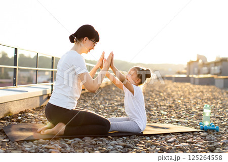 Mother and daughter perform fitness activities on rooftop outdoors, engaging in bonding exercise. Their interaction fosters fitness, movement, and family connection under sunlight. 125264558
