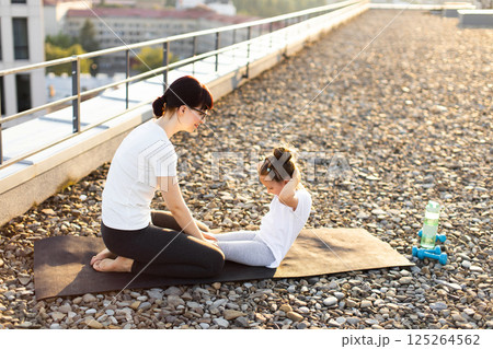 Mother and preteen daughter doing sit-ups on rooftop during sunny day. Woman helps child develop strength and form through guided exercise session. 125264562