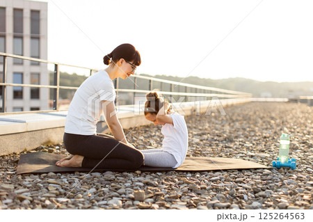 Mother helps young daughter perform abdominal exercises on rooftop during a sunny day, showcasing health and fitness. 125264563