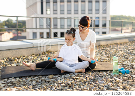 Mother sitting with daughter on rooftop, practicing fitness exercises using small dumbbells, enjoying bonding time. Child learning proper exercise techniques from parent. 125264588
