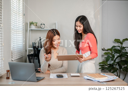 Two women working together in a modern office, discussing documents and using a laptop, showcasing teamwork and collaboration. 125267525