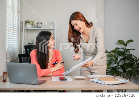 Two women working together in a bright office, discussing documents and using a laptop, showcasing teamwork and productivity. 125267526