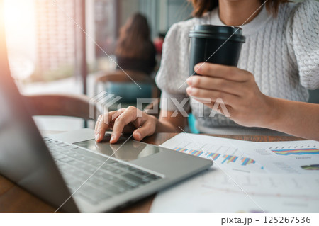 Professional woman multitasking with laptop and coffee, reviewing financial documents in a contemporary office environment. Professional woman multitasking with laptop and coffee, reviewing financial documents in a contemporary office environment. 125267536