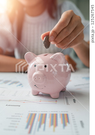Close-up of a hand placing a coin into a pink piggy bank on a table with financial documents and charts. 125267545