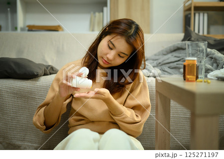 Sick young woman sitting on the floor at home, pouring pills from a bottle into her hand 125271197
