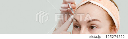 A close-up photograph of a middle-aged woman in which a doctor inserts a syringe into her forehead as part of a rejuvenation and wrinkle smoothing procedure. against the background of a white clinic 125274222