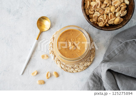 Glass jar with creamy peanut butter on grey background with scattered roasted nuts 125275894