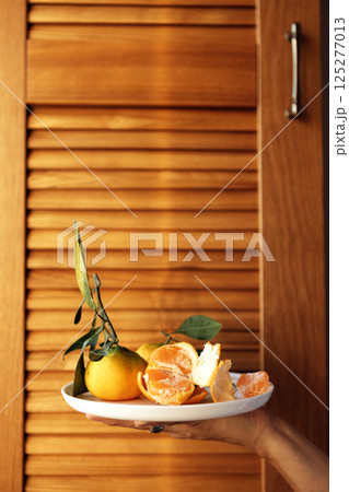 A close-up of a female hand with a ceramic white plate with juicy tangerines. A young woman holds delicious fragrant orange fruits. Healthy and delicious food. 125277013