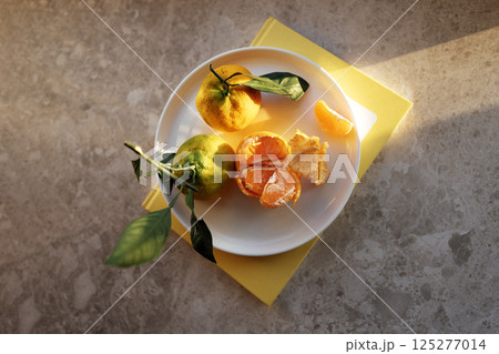 Close-up of juicy tangerines on a ceramic plate. A yellow book and delicious fragrant orange fruits on a marble table. Healthy and delicious food. Beautiful sunlight. 125277014