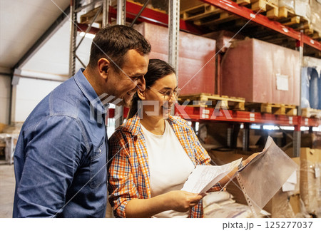 Smiling woman and man in casual clothes are working in warehouse. Young couple of employees laugh and mark presence of product storage in workshop. Smiling woman and man in casual clothes are working in warehouse. Young couple of employees laugh and mark presence of product storage in workshop. 125277037