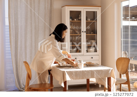 A young cute Asian woman sets the table in the dining room at home. A charming Korean girl sets out plates and cups for a family breakfast. 125277072