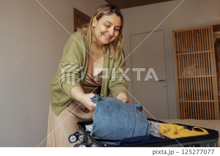 A young female blonde in a beige dress and a green jacket smiles and puts clothes in a suitcase in the bedroom in the house. 125277077
