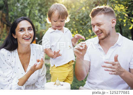 Little cute kid and his parents are tasting festive cake outdoors in garden. Happy family having fun. Little cute kid and his parents are tasting festive cake outdoors in garden. Happy family having fun. 125277081