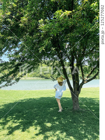Young woman in white clothes and straw hat stands near sprawling tree and looks at river or lake Young woman in white clothes and straw hat stands near sprawling tree and looks at river or lake 125277082