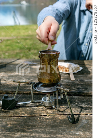 Person brewing traditional Turkish coffee in copper cezve on portable camping stove. Outdoor picnic near lake wooden table surface. Closeup hand stirring coffee. 125278357