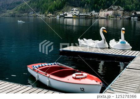 Hallstatt Austria. Wooden dock with swanshaped pedal boats and red motorboat on mountain lake. Background shows picturesque alpine village with traditional houses surrounded by forest. Hallstatt Austria. Wooden dock with swanshaped pedal boats and red motorboat on mountain lake. Background shows picturesque alpine village with traditional houses surrounded by forest. 125278366