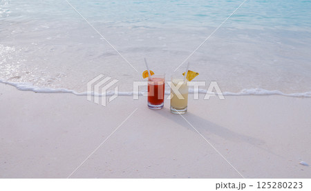 Two colorful delicious cocktails on the ocean. Glasses with cool drinks and pieces of fruit near the clear blue water on the shore. 125280223