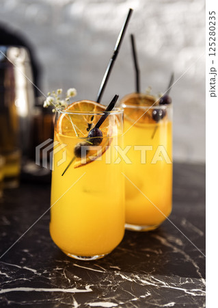 Close-up of two glasses with a citrus cocktail on a black marble table. Tasty chilled drinks are decorated with orange slices, dried flowers and olives. 125280235