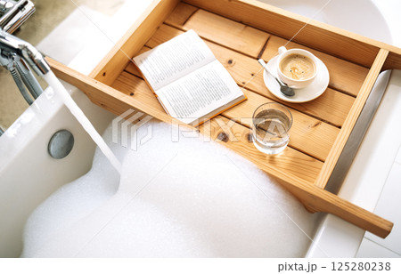 A wooden tray is attached to a bathtub filled with hot fragrant water with fluffy foam. An open book, unbroken coffee and a glass of water on a tray. 125280238