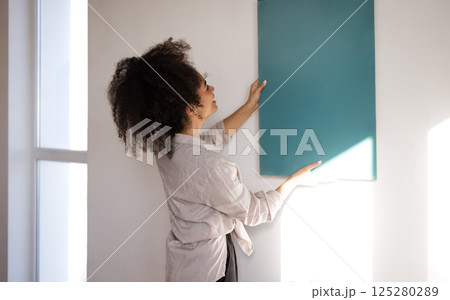 A young beautiful mixed-race girl in casual clothes applies a painted sample to the wall. A cute African woman chooses a paint color. 125280289