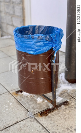 A brown dumpster with a blue trash bag is installed on the sidewalk next to the store. Close-up. 125280633