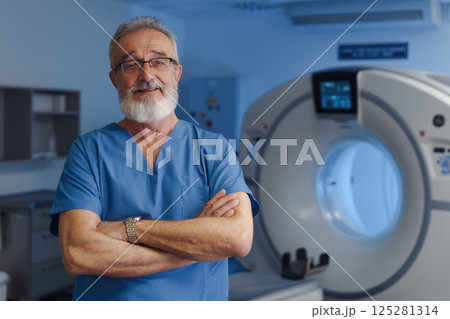 Medical specialist standing by CT scanner, looking at camera, hospital setting. Medical specialist standing by CT scanner, looking at camera, hospital setting. 125281314