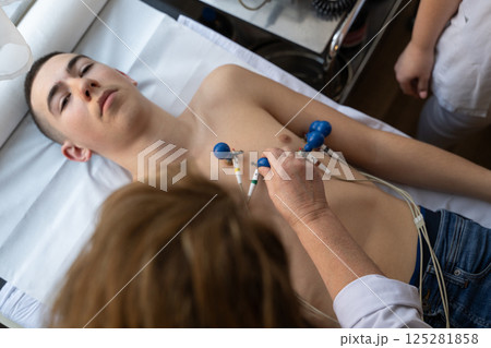 A female doctor performs an ECG on a young man in a hospital room, carefully monitoring his heart health during a routine checkup. 125281858