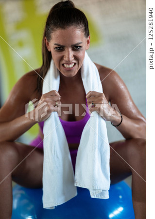 A beautiful and fit woman sitting on a gym ball with a towel, resting and recovering after an intense workout. A beautiful and fit woman sitting on a gym ball with a towel, resting and recovering after an intense workout. 125281940
