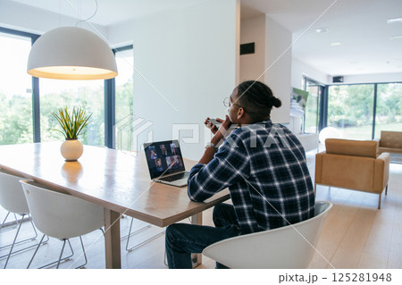 African American businessman in glasses sitting at a table in a modern living room, using a laptop and smartphone for business video chat, conversation with friends and entertainment. African American businessman in glasses sitting at a table in a modern living room, using a laptop and smartphone for business video chat, conversation with friends and entertainment. 125281948