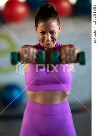 A focused and determined woman performs a strength building dumbbell workout at the gym, showcasing her athletic physique and dedication to fitness. 125281988