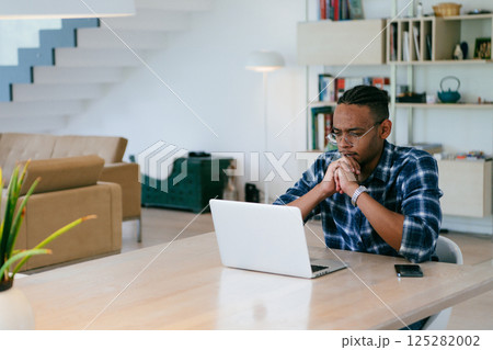 African American influencer in glasses sitting at a table in a modern living room, using a laptop for business video chat, conversation with friends and entertainment. African American influencer in glasses sitting at a table in a modern living room, using a laptop for business video chat, conversation with friends and entertainment. 125282002