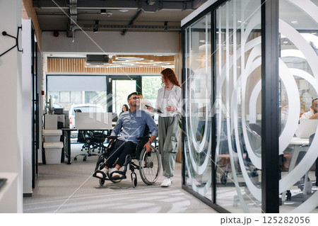 Business colleagues, collaborative business colleagues, including a person in a wheelchair, walk past a modern glass office corridor, illustrating diversity, teamwork and empowerment in the workplace. Business colleagues, collaborative business colleagues, including a person in a wheelchair, walk past a modern glass office corridor, illustrating diversity, teamwork and empowerment in the workplace. 125282056